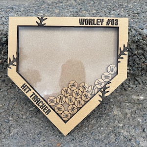 May include: A light brown wooden baseball hit tracker in the shape of home plate. The frame has the text "WORLEY #03" and "HIT TRACKER". Small wooden baseballs are visible inside the frame, with the words "SINGLE", "DOUBLE", and "TRIPLE" on them.