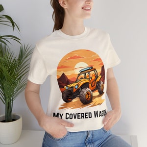 Puede incluir: Camiseta color crema con un dibujo animado de un vehículo todoterreno en un paisaje desértico al atardecer. El texto "MY COVERED WAGON" está impreso debajo. La camiseta la lleva una persona.