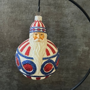 May include: A white glass Christmas ornament with a red and blue striped hat and beard. The ornament features a Santa Claus face with a white beard and a red and blue striped hat. The ornament is decorated with red and blue glitter.