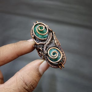 May include: A copper wire wrapped ring featuring two green stones. The stones are set in a spiral design and the ring is crafted from copper wire.