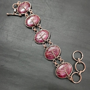 May include: A copper bracelet with five oval, deep red gemstones. Each stone has a wire-wrapped tree design. The bracelet has a toggle clasp and connecting rings. The gemstones are approximately 1.5 cm in length.
