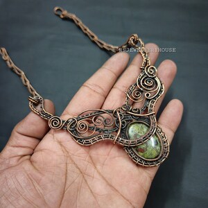 Può includere: Una collana avvolta in filo di rame con un ciondolo in pietra verde e marrone. La collana è su una mano, e il testo "RSJEWELLERYHOUSE" è visibile sullo sfondo.
