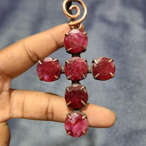 Puede incluir: Un colgante de cruz artesanal con grandes piedras preciosas facetadas de color rojo intenso engastadas en un marco de metal oscuro. La cruz tiene una espiral de color cobre en la parte superior para colgar. Las piedras son de un color vibrante.