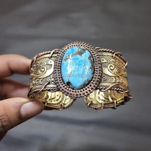 May include: A handcrafted cuff bracelet featuring a large, oval turquoise stone set in a copper wire frame. The wide, ornate band is made of gold-toned metal with embossed bird and floral designs. The bracelet is held by a hand.