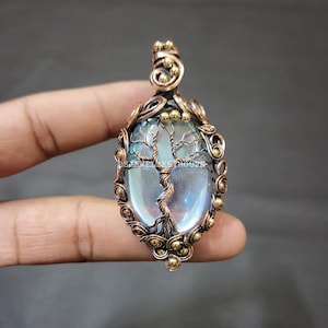 May include: An oval pendant featuring a wire-wrapped design with a tree motif. The pendant has a central iridescent stone, framed by copper-colored wire and accented with small gold-colored beads. The overall design is intricate and detailed.