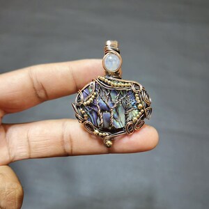 May include: A handcrafted pendant with a wire-wrapped design, abalone shell, and a moonstone. The pendant has a copper-coloured wire frame with gold accents and a tree design. The pendant is held in a hand.