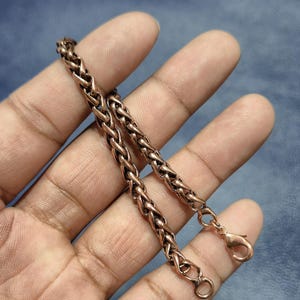 以下が含まれることがあります： 小麦のリンクデザインの銅色のチェーンブレスレット。ブレスレットは手で持たれており、その複雑な織り模様とロブスタークラスプの留め金を示しています。温かみのある金属の色合いは、青い背景と対照的です。