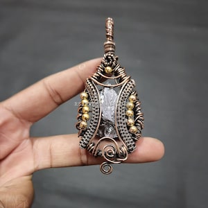 Może przedstawiać: Ręcznie robiony wisiorek z przezroczystym, fasetowanym kamieniem szlachetnym, zamkniętym w skomplikowanym drucie miedzianym. Praca z drutu jest szczegółowa z koralikami w kolorze złotym, tworząc unikalny i artystyczny design. Wisiorek jest zawieszony na pętelce.