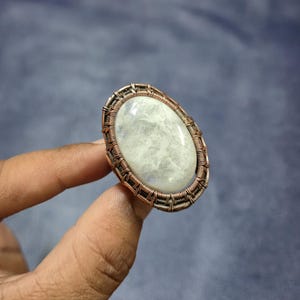 May include: Oval-shaped ring featuring a large, pale gemstone set within a copper wire frame. The gemstone has a milky, translucent appearance. The ring is held against a muted blue background, showcasing the handcrafted jewelry.