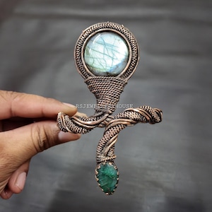 Peut inclure: Un bracelet en fil de cuivre enroulé avec une grande pierre précieuse ronde en labradorite et une petite pierre précieuse en émeraude au bas.