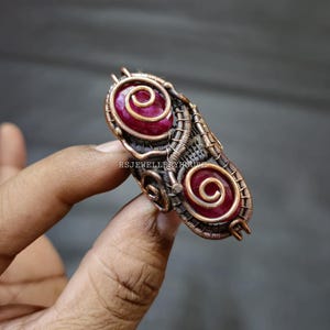 Puede incluir: Un anillo envuelto en alambre de cobre con dos piedras preciosas rojas. Las piedras preciosas están engastadas en espiral y el anillo está hecho con un diseño delicado de alambre.
