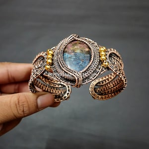 Peut inclure: Un bracelet en fil de cuivre enroulé avec une grande pierre précieuse multi-colore de forme ovale sertie au centre. Le bracelet est accentué de petites perles dorées.