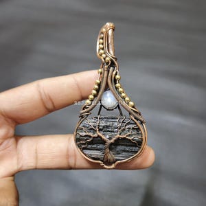 May include: A handcrafted pendant featuring a wire-wrapped design. The pendant showcases a black stone with a wire tree of life design, accented with a small, round, light-colored stone. The pendant is made with copper-colored wire and gold-colored beads.