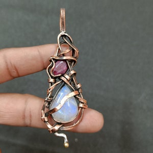 以下が含まれることがあります： 涙型のムーンストーンと小さな赤い宝石を特徴とする、銅線で巻かれたペンダント。ペンダントには金色のベイルが付いています。