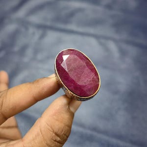 May include: A large, oval-cut ruby gemstone ring. The ruby is a deep, rich red color, set in a gold-toned metal band with an ornate, textured design. The ring is held up by a hand, showcasing the size and brilliance of the stone.