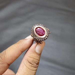 May include: A copper-coloured ring with an oval, deep red gemstone. The ring's setting has an ornate, textured design around the gemstone. The ring is held up against a grey background.