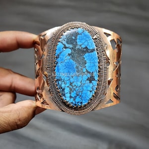 Puede incluir: Un brazalete de cobre con una gran piedra turquesa ovalada engastada en el centro. La piedra tiene un color azul y verde con un patrón veteado. El brazalete tiene un delicado diseño calado alrededor de la piedra.