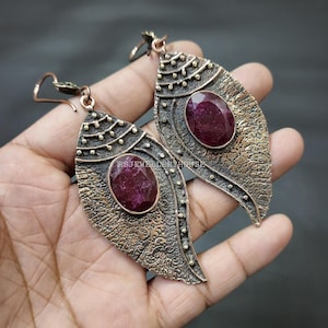 May include: A pair of dangle earrings with a leaf-shaped design. The earrings are made of copper and feature a deep red gemstone in the center of each leaf. The leaves have a textured surface and are decorated with small gold dots.