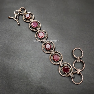 Puede incluir: Una pulsera de estilo vintage con seis grandes piedras preciosas redondas de color rojo intenso engastadas en marcos circulares ornamentados. La pulsera tiene un metal plateado con un diseño de cuerda trenzada y un cierre de broche. El texto "RSJEWELRYHOUSE" es visible.