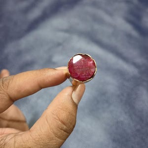 May include: A ruby ring with a large, faceted, oval-shaped red gemstone. The gemstone is set in a gold-colored bezel on a silver band. The ring is held up against a blue background.