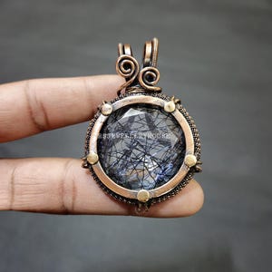May include: A handcrafted pendant featuring a large, round, faceted gemstone with black inclusions, set in a copper-coloured metal frame. The frame has decorative wirework and small gold-coloured accents. The pendant is held by a hand against a dark background.