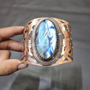 Puede incluir: Brazalete de puño de cobre con una gran piedra ovalada de labradorita. La piedra muestra iridiscencia azul y verde. El brazalete tiene un diseño floral calado y un borde texturizado.