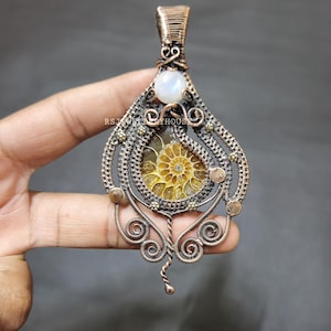 May include: A copper wire wrapped pendant featuring a golden ammonite fossil and a white moonstone. The pendant is intricately designed with swirls and loops.