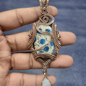 Può includere: Ciondolo artigianale in filo di rame con una pietra maculata con punti blu. La pietra è racchiusa in un'intricata filigrana, con una pietra a forma di goccia che pende sotto. Il ciondolo è tenuto in una mano.