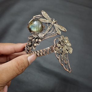 May include: A copper-toned bracelet featuring a dragonfly and floral design. The bracelet has a large, round, iridescent stone and is held by a hand against a dark background. The bracelet's design includes a dragonfly, flowers, and leaves.