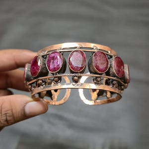 Puede incluir: Un brazalete de puño de color cobre con piedras preciosas rojas oscuras de corte ovalado. El brazalete tiene un diseño abierto con elementos decorativos de metal y está sujeto por una mano.