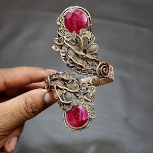 Pode incluir: Um bracelete envolto em fio de cobre com duas pedras de rubi vermelho engastadas no design. O bracelete apresenta um design floral com detalhes intrincados.