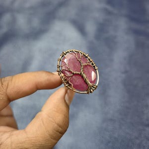 Puede incluir: Anillo ovalado con una gran gema facetada de color rubí. La piedra está engastada en un diseño de alambre color cobre que se asemeja a un árbol. El anillo se sostiene sobre un fondo azul.