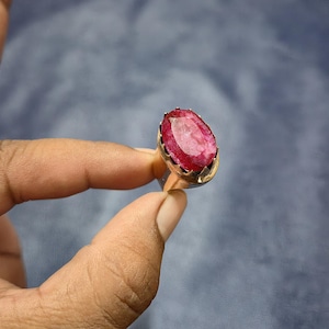 May include: A gold-toned ring featuring a large, oval-cut ruby gemstone. The ruby is a vibrant red color, set in a detailed, ornate bezel. The ring is held up against a muted blue background.