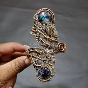 Peut inclure: Un bracelet en fil de cuivre enroulé avec deux cabochons en verre dichroïque bleu et violet. Le bracelet présente un motif floral avec un travail de fil complexe.