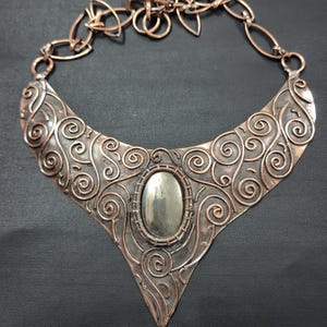May include: A copper-toned statement necklace with an oval, silver-colored stone at the center. The necklace features intricate scrollwork and a chain with decorative links. The pendant has a triangular bottom.