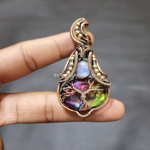 May include: A handcrafted copper pendant with a colorful gemstone and wire-wrapped design. The pendant features a tree of life motif, a small moonstone, and gold-colored beads. The gemstone displays shades of purple, green, and yellow.