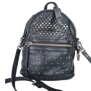 Puede incluir: Una mochila de cuero negro con una superficie texturizada adornada con numerosos pequeños remaches metálicos de color plateado. El bolso incluye un asa superior, un bolsillo frontal con cremallera plateada y correas ajustables con clips metálicos.