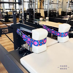 Puede incluir: Máquina de reformer Pilates con reposacabezas y asientos acolchados blancos. Los reposacabezas tienen correas estampadas con diseños geométricos azules, rosas y blancos. La máquina está en un gimnasio.