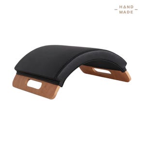 May include: A black Pilates arc with wooden handles. The arc is curved and upholstered in black material. The wooden handles are light brown. The text "HAND MADE" is in the upper right corner.