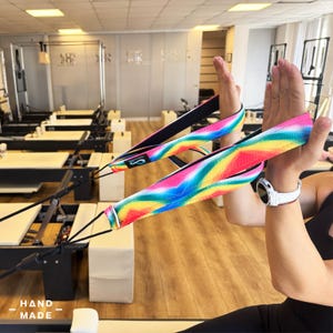 Puede incluir: Correas de reformer de Pilates con un vibrante estampado tie-dye arcoíris. Las correas son sostenidas por una persona en un estudio de Pilates. Las correas tienen un respaldo negro y están conectadas a la máquina reformer con cuerdas negras. Las palabras "HAND - MADE" están en la parte inferior.