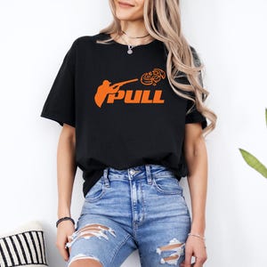 May include: Black t-shirt with the word "PULL" in orange, accompanied by an orange graphic of a person aiming a shotgun. The shirt is a casual, oversized fit.
