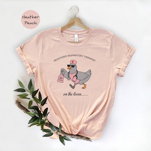 May include: Heather peach-colored t-shirt featuring a cartoon duck dressed as a respiratory therapist. The duck wears a nurse's cap, sunglasses, and a stethoscope, holding an oxygen tank. Text reads "REGISTERED RESPIRATORY THERAPIST" and "on the loose....."