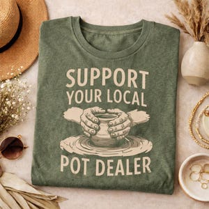 May include: Green t-shirt with the text "SUPPORT YOUR LOCAL POT DEALER" in a vintage style. The design features hands shaping a pot on a pottery wheel. The shirt is made of a soft, comfortable material.