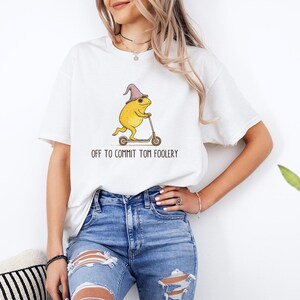 May include: White t-shirt featuring a yellow frog wearing a purple wizard hat, riding a scooter. The text "OFF TO COMMIT TOM FOOLERY" is printed below the frog. The shirt is paired with blue jeans.