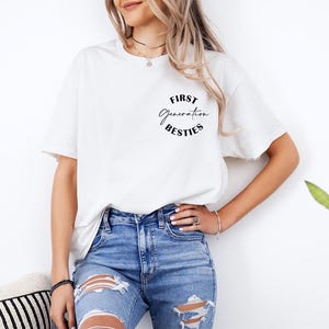 May include: White t-shirt with the text "FIRST Generation BESTIES" in a black, circular design. The shirt is paired with blue denim jeans. The image is taken in a well-lit setting.