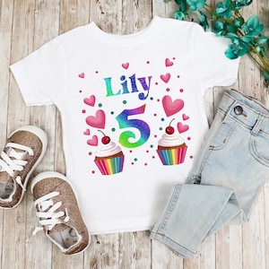 May include: White t-shirt with a rainbow glitter number 5, the name "Lily" and cupcake graphics. Pink hearts are scattered around the design.
