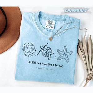 May include: A light blue t-shirt featuring a black line art graphic of a seashell with flowers, a sea turtle, and a starfish. Below the graphic, the text reads "Be Still And Know That I Am God" and "PSALM 46:10". The shirt has a Comfort Colors label.