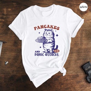 May include: White t-shirt featuring a cartoon cat holding a plate of pancakes. The text "Pancakes and panic attacks" is printed on the shirt.