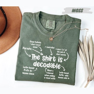 May include: An olive green t-shirt with white text that reads The shirt is decodable, surrounded by linguistic notes including Closed Syllable, Digraphs, r-controlled, s says /z/, Latin-based Prefix, Silent-e, and Latin-based Suffix, with a gold necklace.