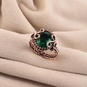 May include: A ring featuring a large, oval-cut dark green gemstone set within a complex, hand-wrapped copper wire frame. The metal has an oxidized finish, highlighting the intricate swirls and coiled wire details surrounding the central stone.